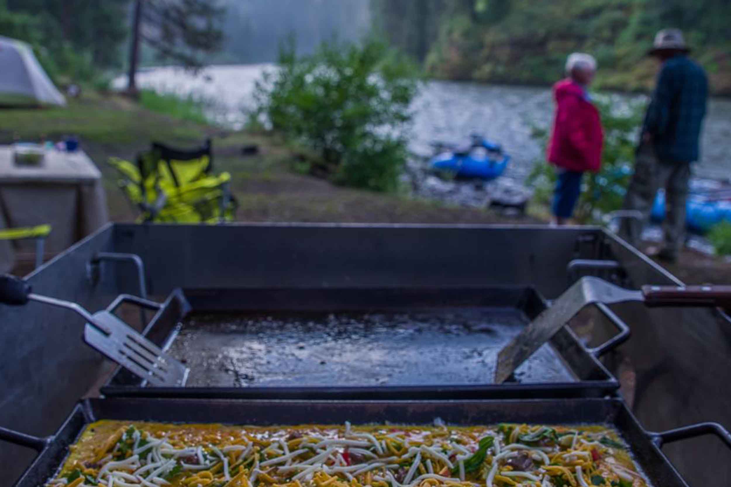 Camp griddle of vegetables on the riverbank, guests at the water behind